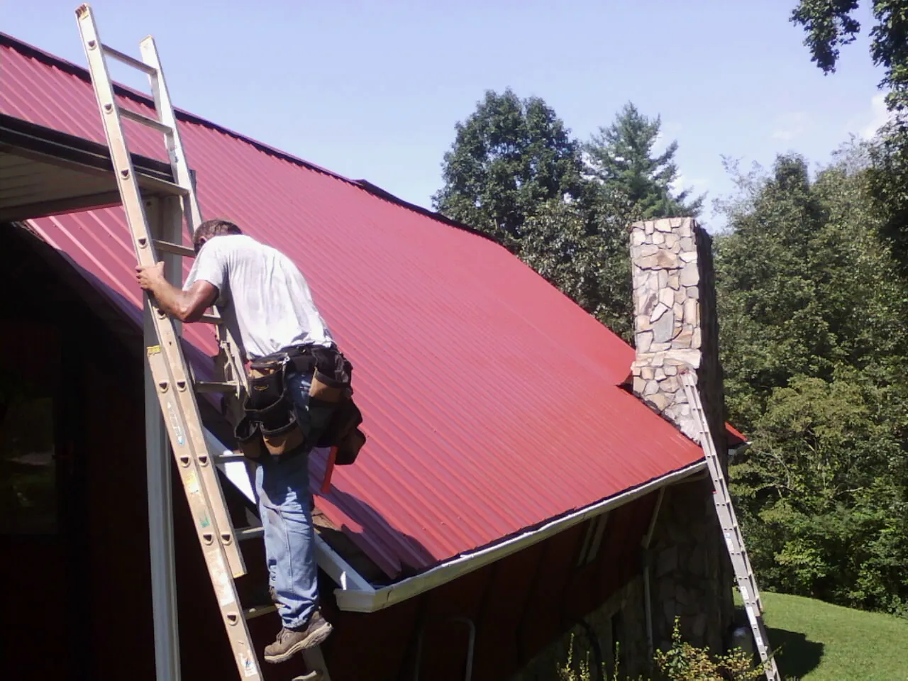 Roofer on a ladder beside a red metal roof and stone chimney — Soffit & Fascia Repair by Roofing Brothers in Capitol Heights